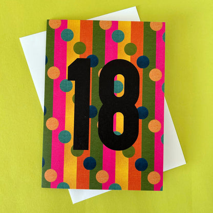 A colourful 18th birthday greeting card with bold black numerals '18' over vertical stripes of pink, orange, yellow, green, and blue polka dots, shown with a white envelope on a bright yellow-green background.