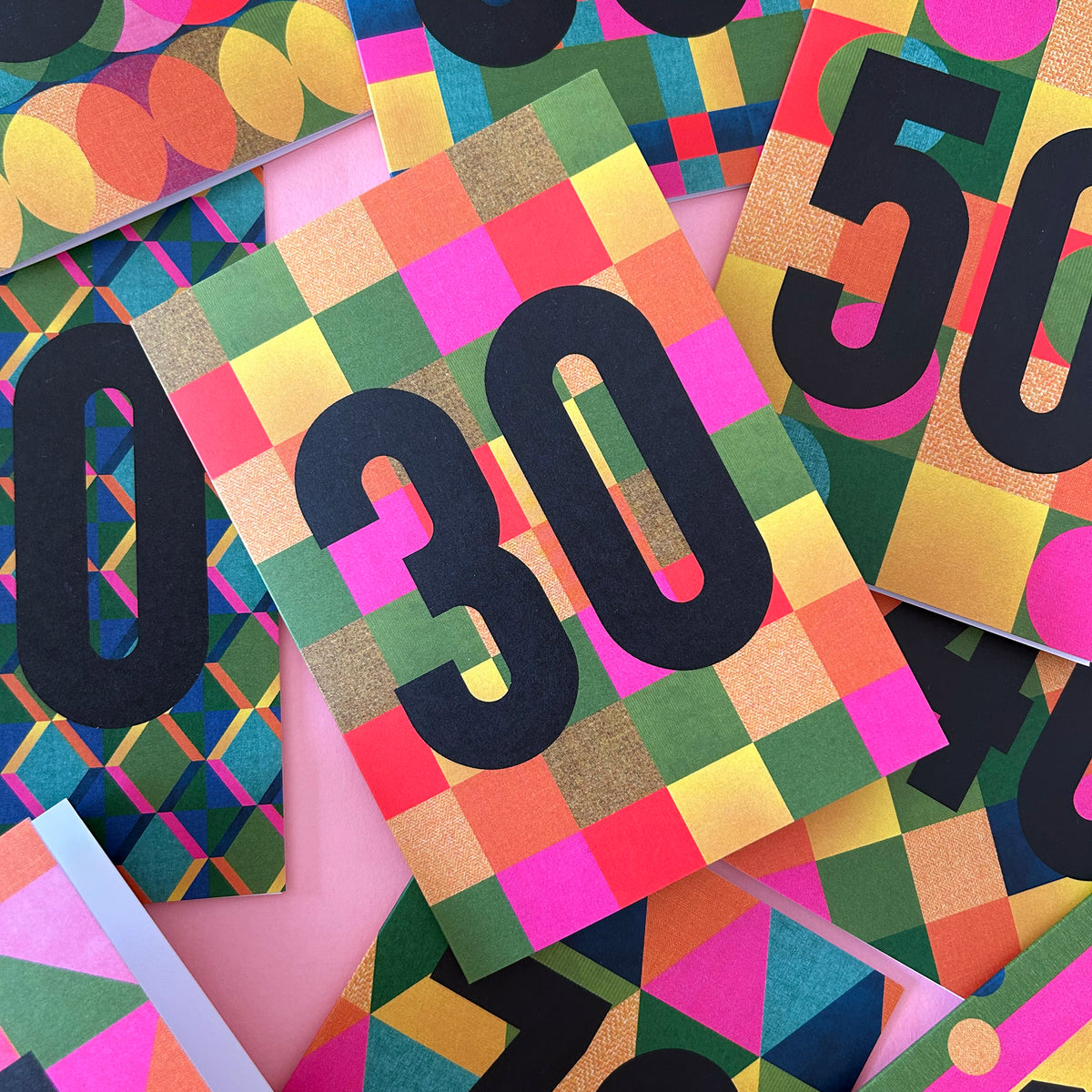 A close-up, top-down view of overlapping colourful patterned cards or envelopes featuring large black numbers, including 30 and 50, on a bright multicolour background.