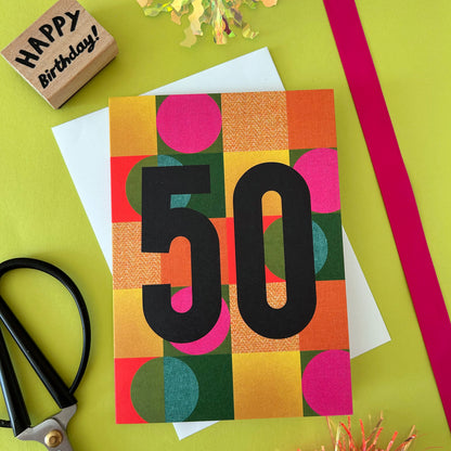 A colourful 50th birthday greeting card with a bold black '50' over a patchwork of orange, pink, green, and yellow geometric shapes, styled on a bright yellow-green background with scissors, a wooden stamp, and ribbon.
