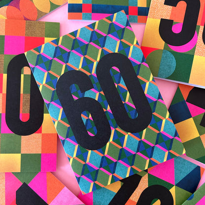 A close-up flat lay of colourful patterned greetings cards with large black numbers The visible central item has a geometric multicolour design in blue, green, pink, yellow, and orange on a pink background.