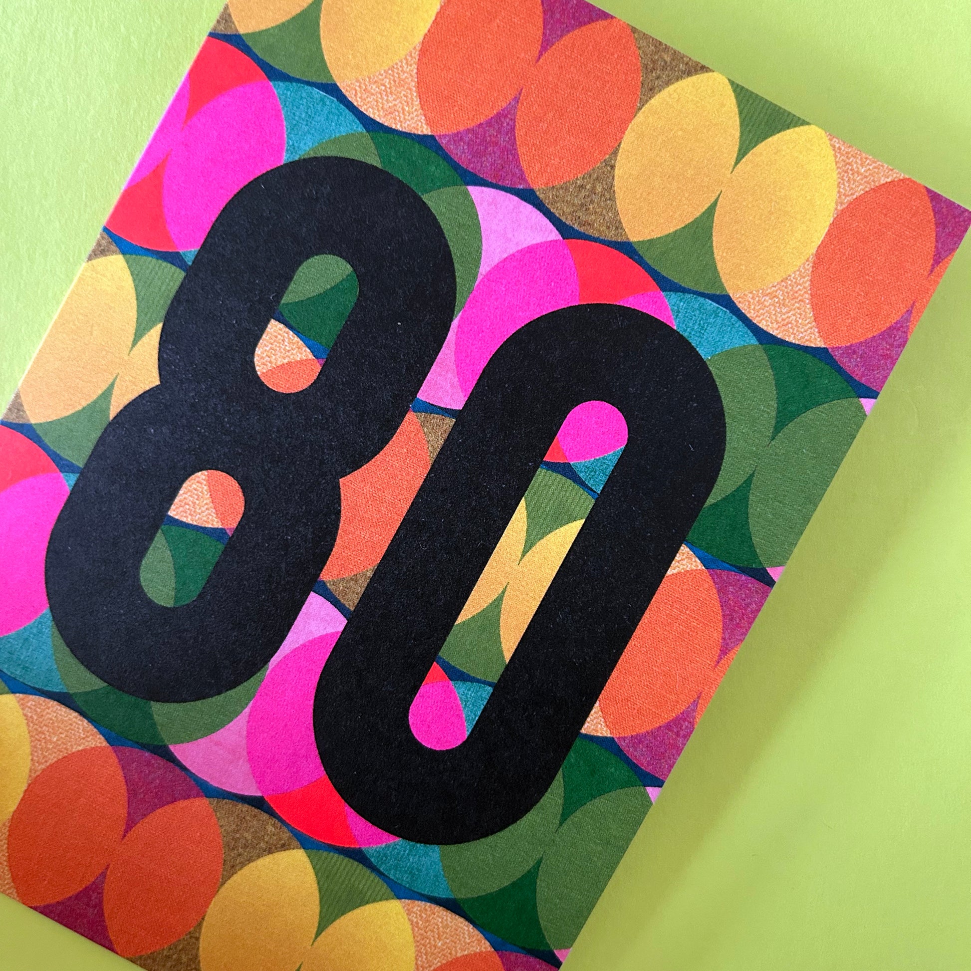 A bright greeting card with a colourful retro pattern of overlapping circles in pink, yellow, orange, green, and blue, featuring a large black '80' printed on the front, set against a light green background.
