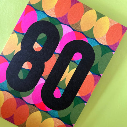A bright greeting card with a colourful retro pattern of overlapping circles in pink, yellow, orange, green, and blue, featuring a large black '80' printed on the front, set against a light green background.