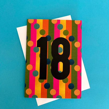 A colourful birthday card featuring vertical stripes in orange, yellow, green, pink, and blue with large black '18' text, shown with a white envelope on a bright blue background.