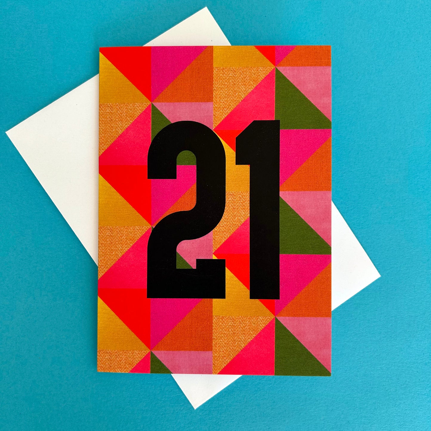 A square greeting card with a colourful geometric triangle pattern in pink, orange, yellow, and green, featuring the number '21' in large black font. The card is displayed on top of a white envelope against a solid blue background.