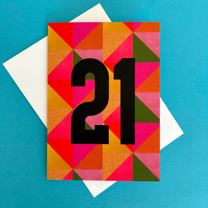 A square greeting card with a colourful geometric triangle pattern in pink, orange, yellow, and green, featuring the number '21' in large black font. The card is displayed on top of a white envelope against a solid blue background.