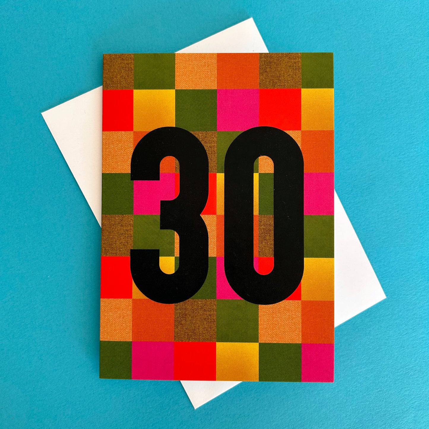 A square greeting card with a colourful mosaic grid pattern in orange, green, pink, red, and yellow, featuring the number '30' in bold black font. The card is displayed on top of a white envelope against a solid bright blue background.