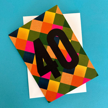 A bright greeting card with a colourful geometric checkered pattern in orange, yellow, green, pink, and navy, featuring a large black '40' printed on the front. A plain white card is partially visible underneath, set against a solid bright blue background.