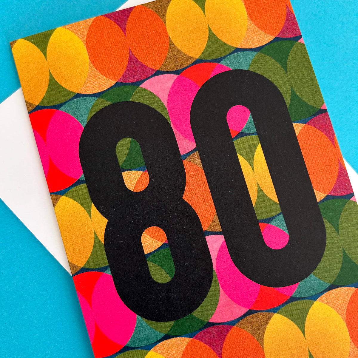 A bright greeting card with a bold black '80' printed over a colourful retro pattern of overlapping circles in orange, yellow, pink, green, and blue, shown on a bright blue background with a white envelope partially visible.