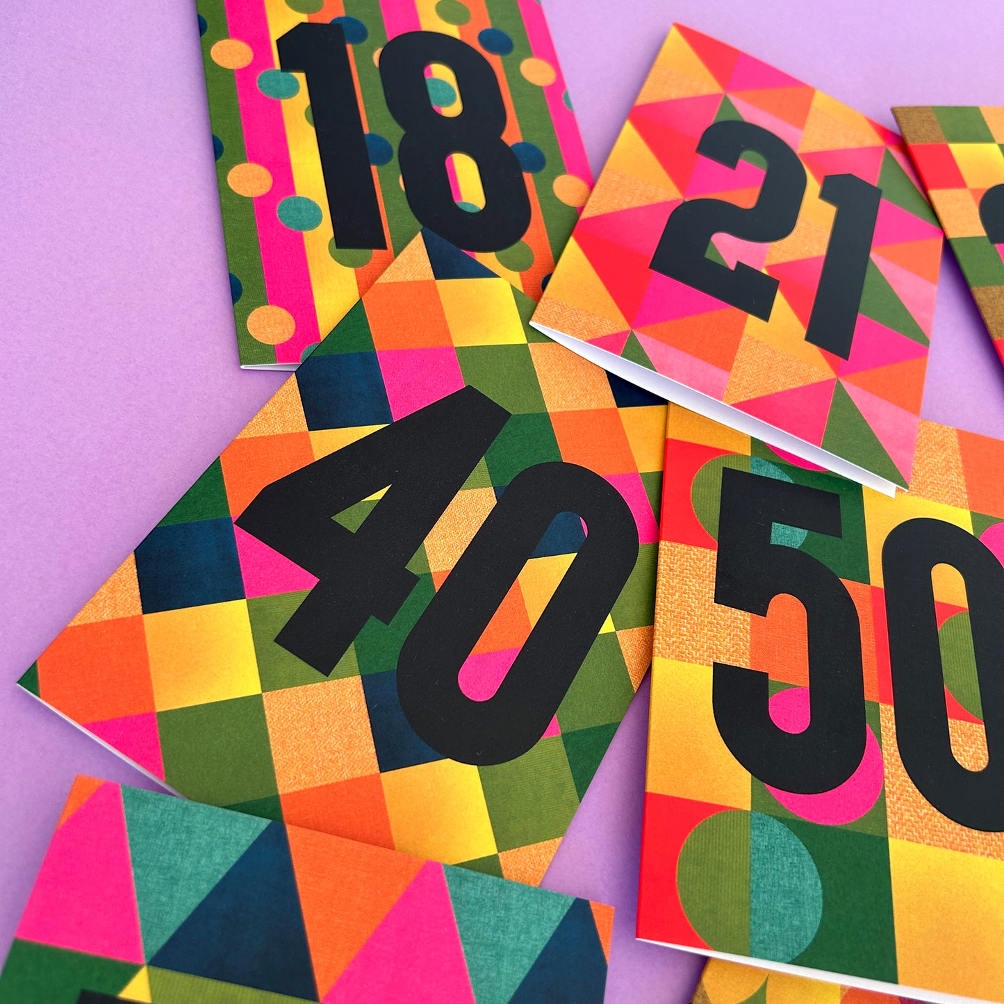 A close-up of several colourful printed cards or book covers arranged on a lavender background, each featuring large black numbers (18, 21, 40, 50) over a bright geometric pattern of pink, orange, yellow, green, and blue shapes.