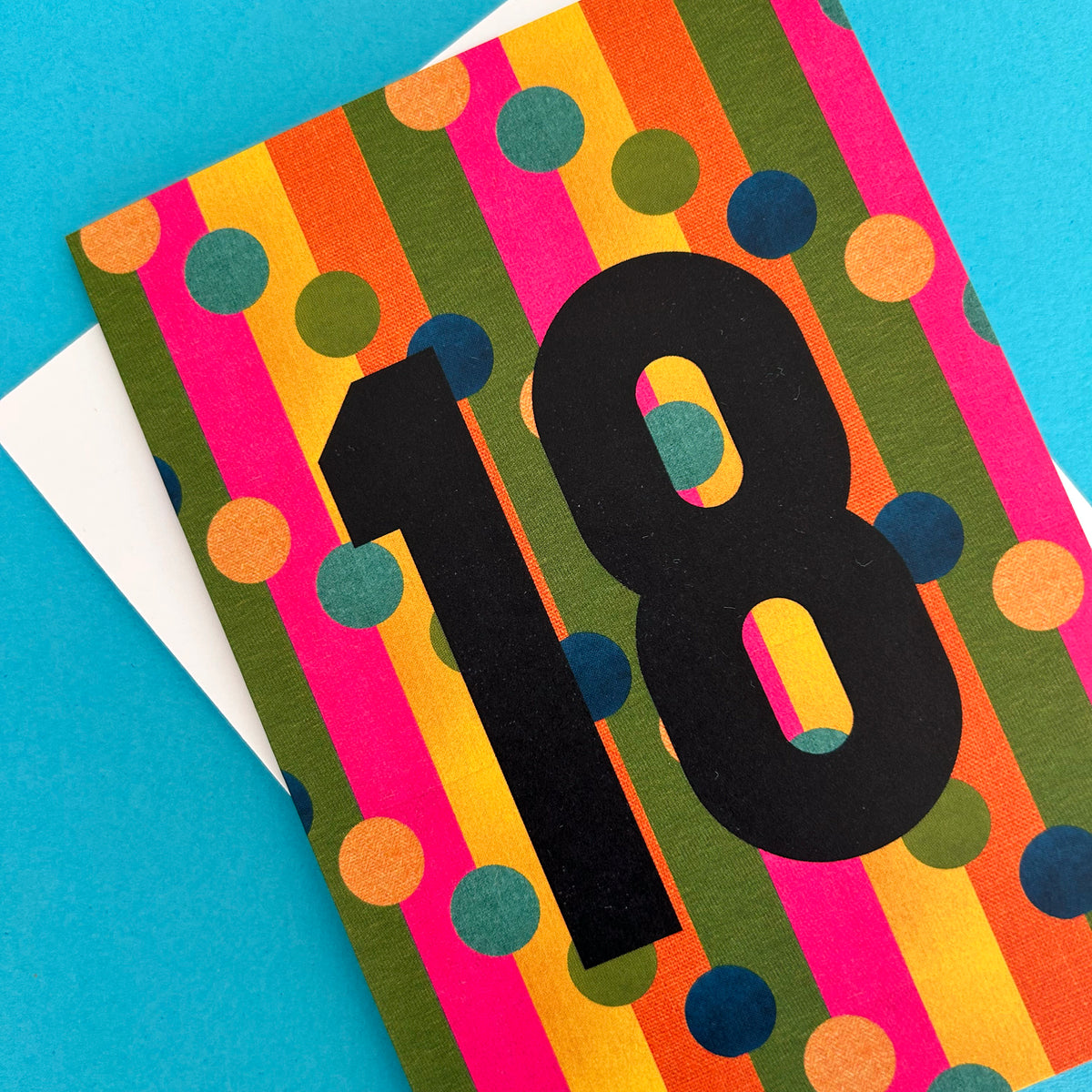 A colourful birthday card with vertical stripes in pink, orange, yellow, olive green, and blue, overlaid with a large black number 18. A white envelope is partially visible underneath on a bright blue background.