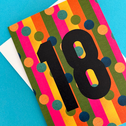 A colourful birthday card with vertical stripes in pink, orange, yellow, olive green, and blue, overlaid with a large black number 18. A white envelope is partially visible underneath on a bright blue background.