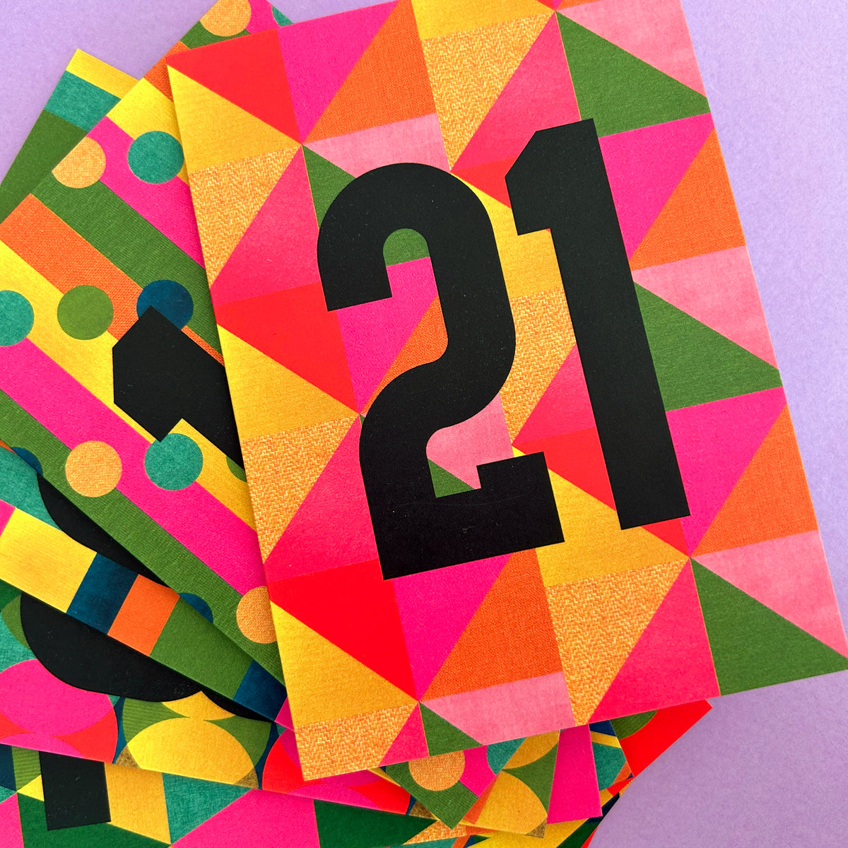 A stack of colourful geometric paper cards or envelopes on a light purple background. The top card prominently displays the black number 21 over a bright multicolour pattern of pink, orange, yellow, green, and red triangles.