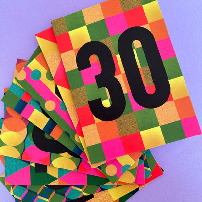 A stack of colourful square cards or paper covers fanned out on a light purple background. The top card features a bold black number 30 over a multicolour checkerboard and geometric pattern in green, yellow, orange, pink, and blue.