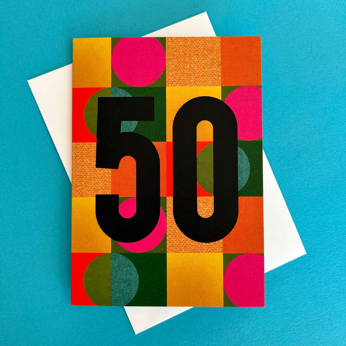 A bright greeting card with a colourful geometric pattern of pink circles, orange and yellow blocks, and green textured squares, featuring the number '50' in bold black text. A white envelope is partially visible behind it on a bright blue background.