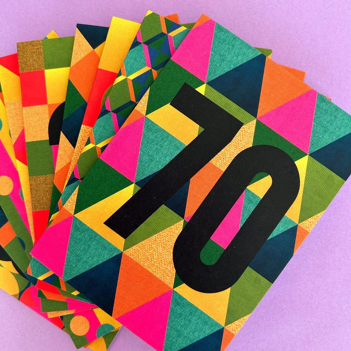 A stack of colourful geometric patterned cards or paper pieces fanned out on a light purple background. The top card features a bold black number 70 over a multicolor triangle pattern in pink, green, yellow, orange, and blue.