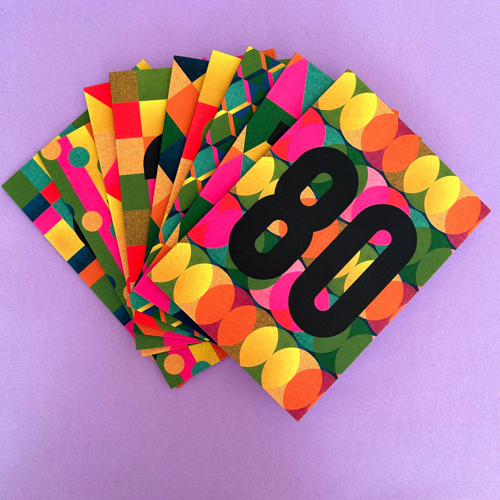 A stack of colourful cards fanned out on a light purple background. The front card has a bold black number 80 over a retro pattern of pink, yellow, green, orange, and blue circles and blocks.