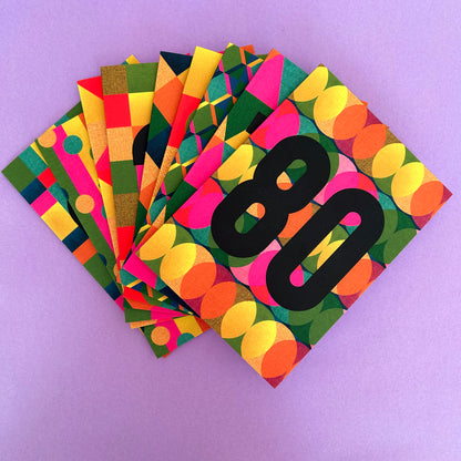 A stack of colourful cards fanned out on a light purple background. The front card has a bold black number 80 over a retro pattern of pink, yellow, green, orange, and blue circles and blocks.