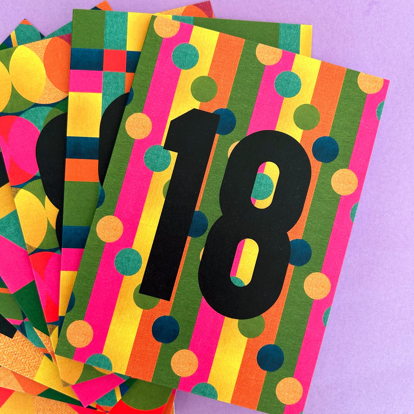 A close-up of a colourful striped and polka-dot patterned card or ticket with the number 18 in large black font, layered over similar cards on a light purple background.