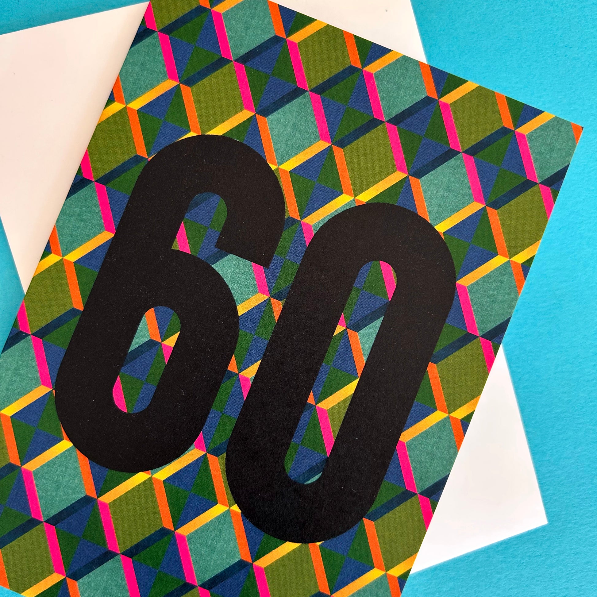 A bright greeting card with a bold black '60' printed over a colourful geometric pattern of green, blue, pink, and orange triangles, layered on top of a white envelope against a bright blue background.