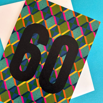A bright greeting card with a bold black '60' printed over a colourful geometric pattern of green, blue, pink, and orange triangles, layered on top of a white envelope against a bright blue background.