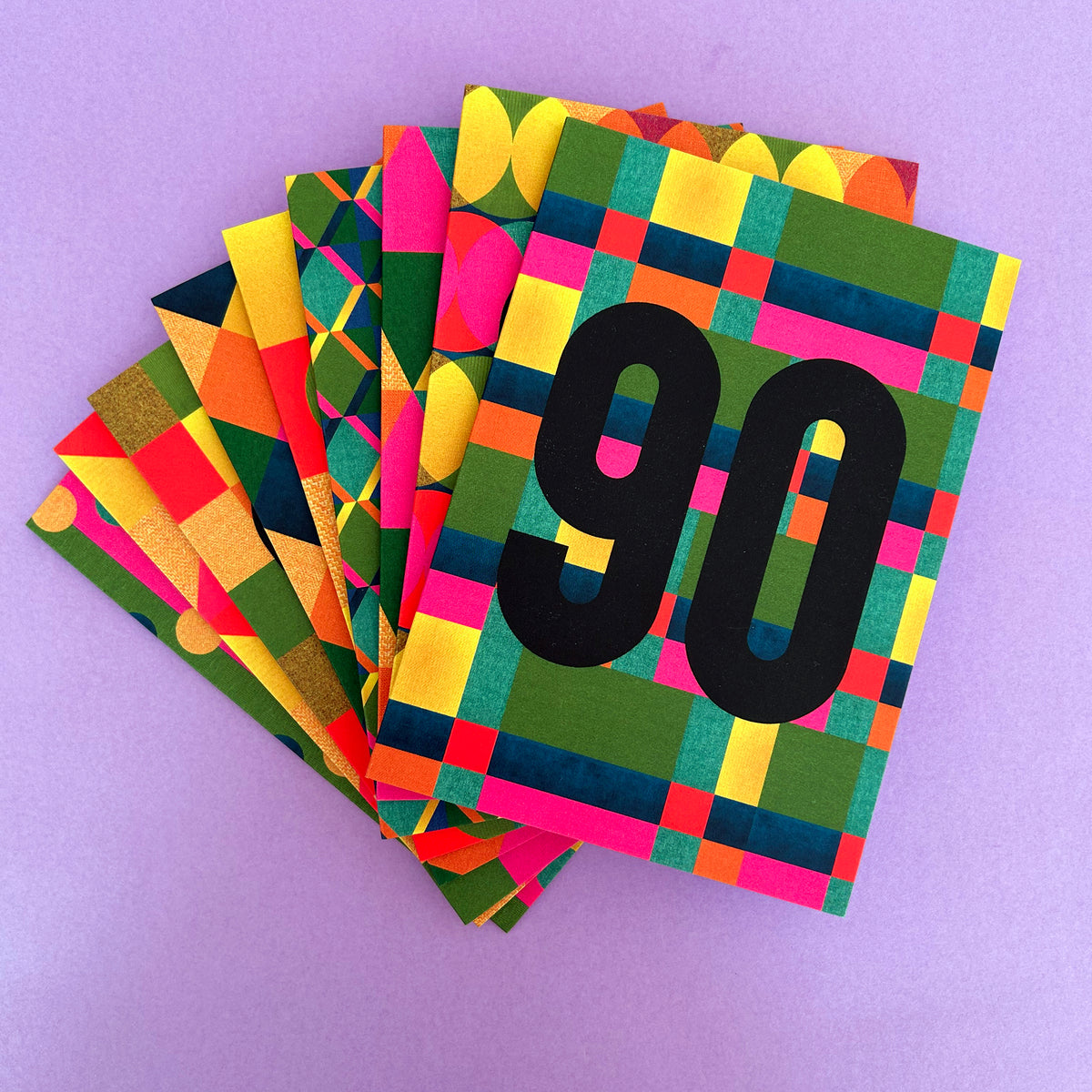 A stack of colourful patterned cards fanned out on a light purple background. The top card features a bold black '90' over a green, yellow, pink, and blue geometric plaid pattern.