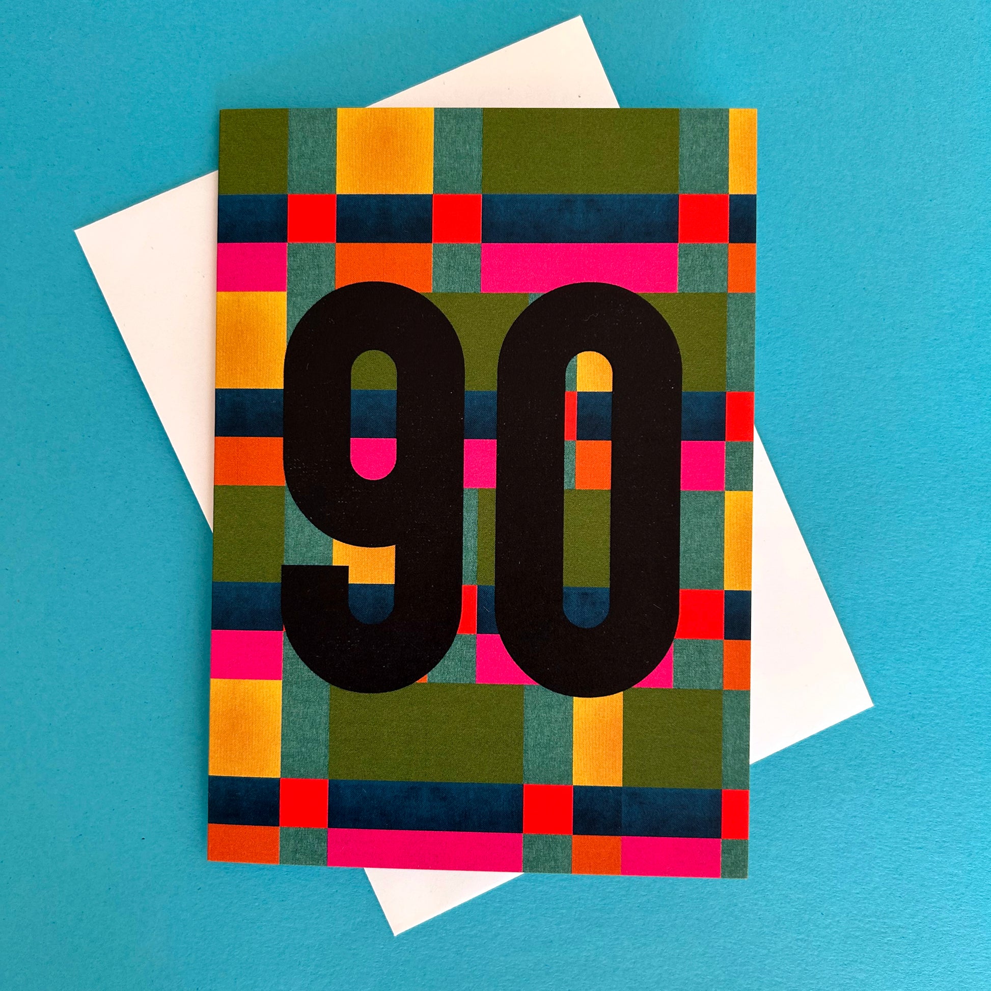 A bright greeting card with a colourful geometric pattern in green, yellow, orange, pink, and blue, featuring the number '90' in large black letters. A white envelope is partially visible behind the card on a bright blue background.
