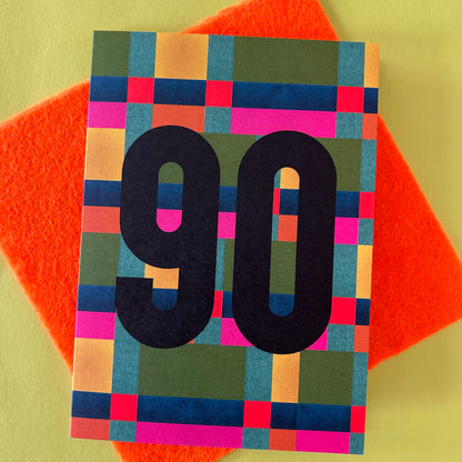 A bright greeting card with a colourful retro pixelated block pattern and the number '90' in large black font, placed on top of an orange felt square over a pale yellow background.