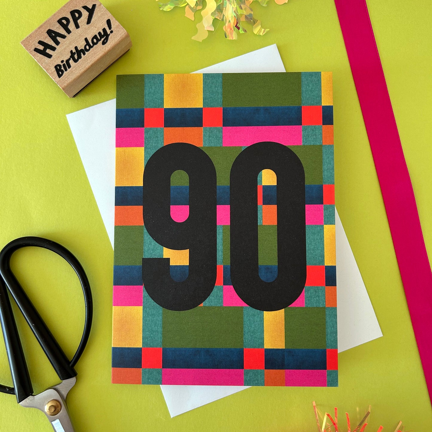 A colourful 90th birthday card with a geometric block pattern in green, blue, pink, orange, and yellow, featuring a large black '90' in the centre. The card is styled on a bright yellow-green background with craft scissors, a wooden 'Happy Birthday!' stamp, white paper, and a pink ribbon.
