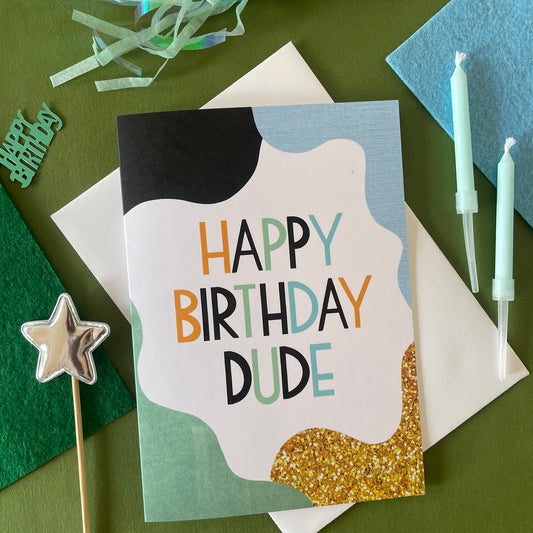 Image shows a white greetings crad with pale blue, green, black and gold abstract shapes and the words 'Happy Birthday Dude'
