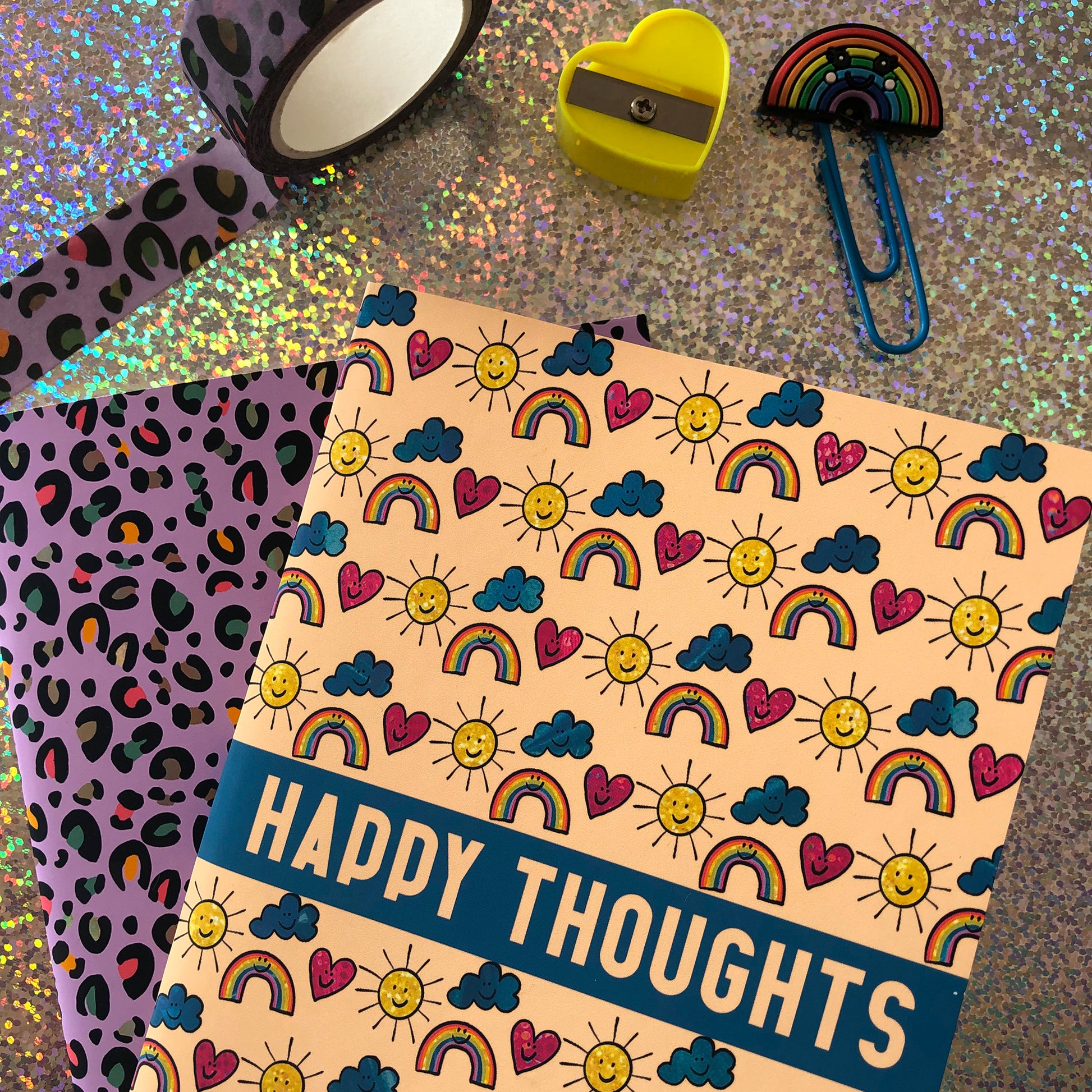 Image shows a patterned notebook with illustrations of suns, clouds, hearts and rainbows with smiling faces. The notebook says Happy thoughts on the cover
