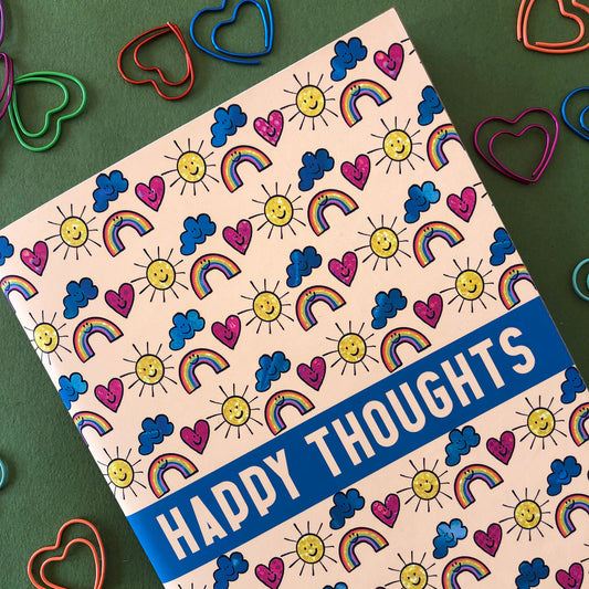 Image shows a patterned notebook with illustrations of suns, clouds, hearts and rainbows with smiling faces. The notebook says Happy thoughts on the cover