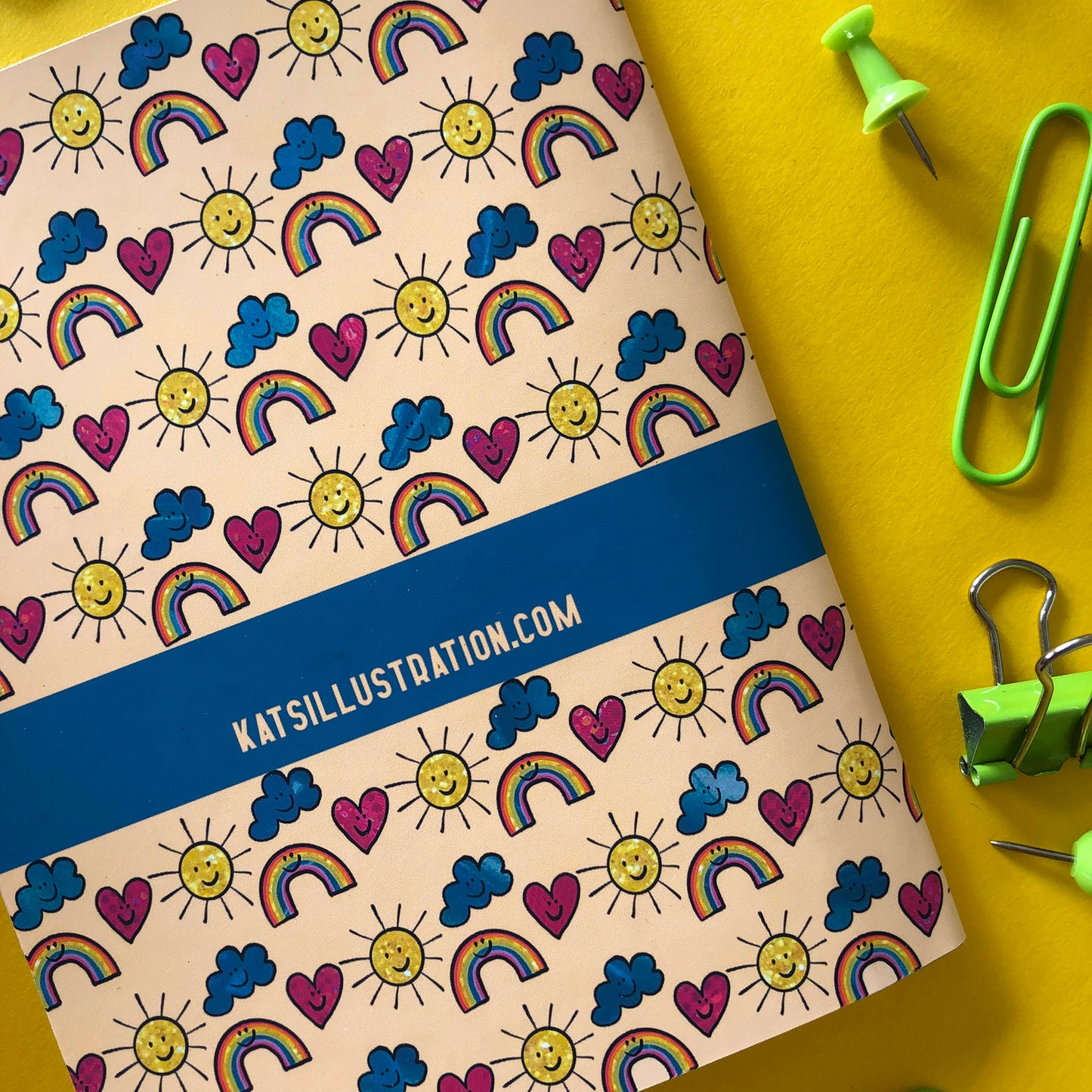 Image shows the back of a patterned notebook with illustrations of suns, clouds, hearts and rainbows with smiling faces.