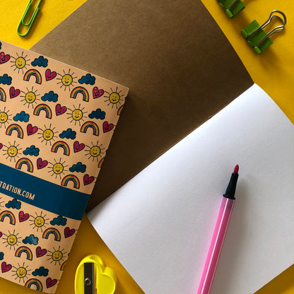 Image shows the back of a patterned notebook with illustrations of suns, clouds, hearts and rainbows with smiling faces and a n open notebook with a kraft inside cover and blank pages.
