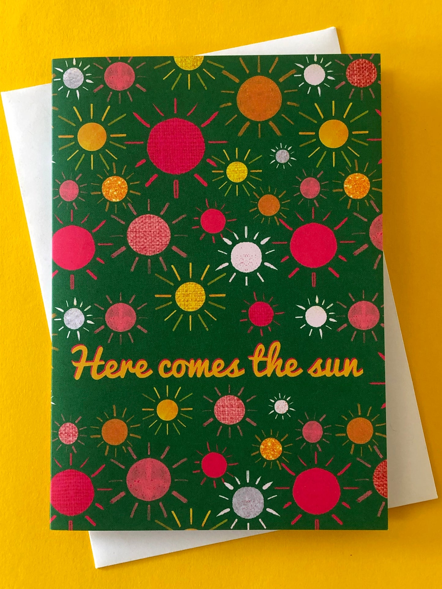 Green everyday greetings card that says 'here comes the sun' and features a fun pattern of colourful suns