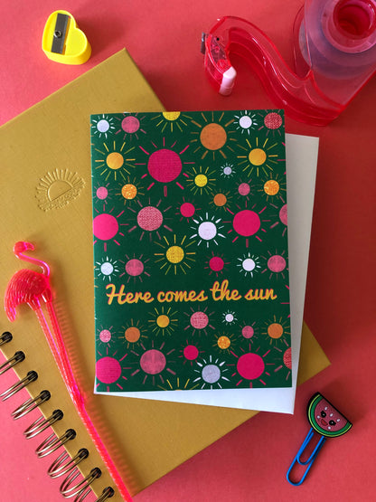 Green everyday greetings card that says 'here comes the sun' and features a fun pattern of colourful suns
