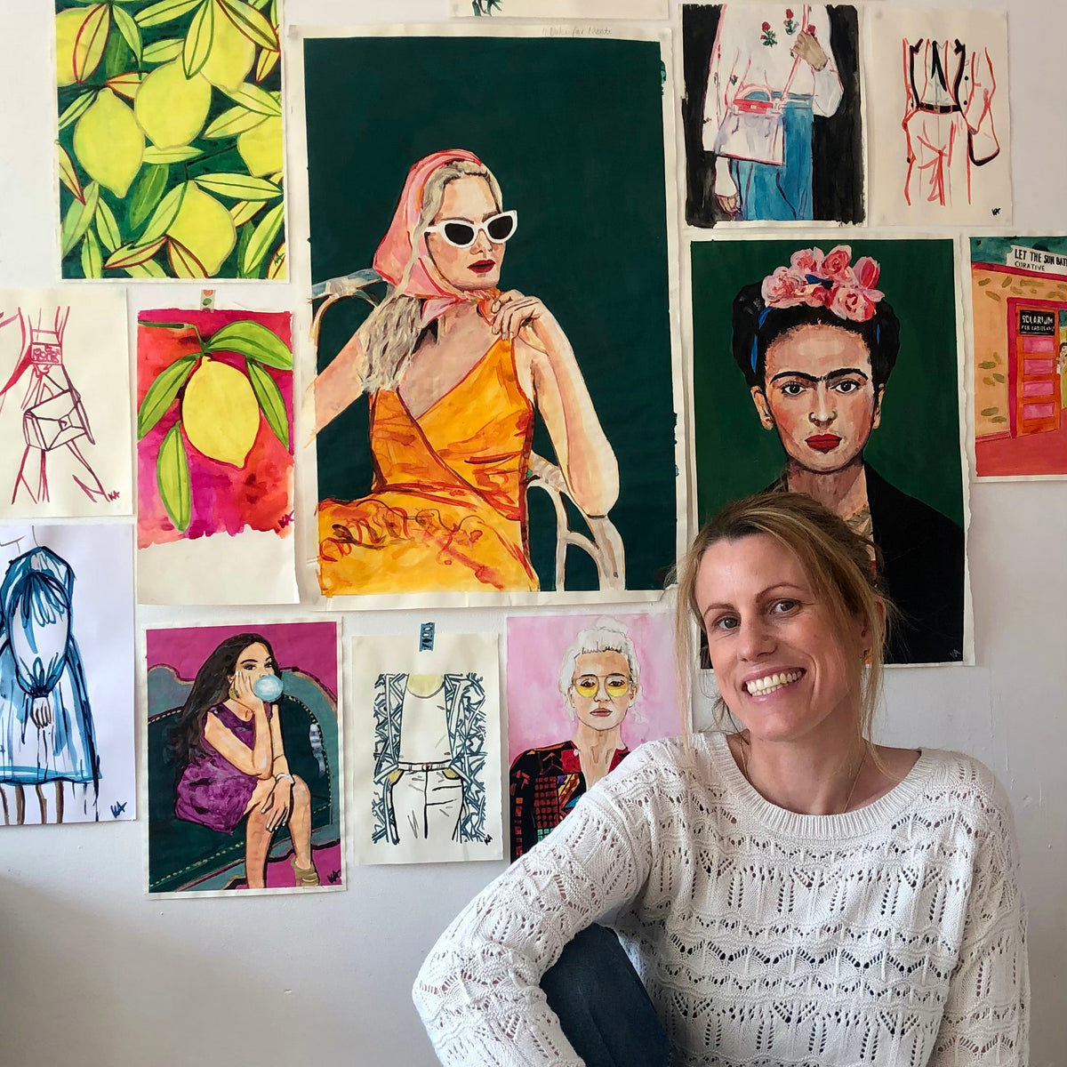 A smiling woman with blonde hair sits in a room, wearing a white knit sweater and blue jeans, posed in front of a wall covered with colourful fashion illustration prints and sketches.