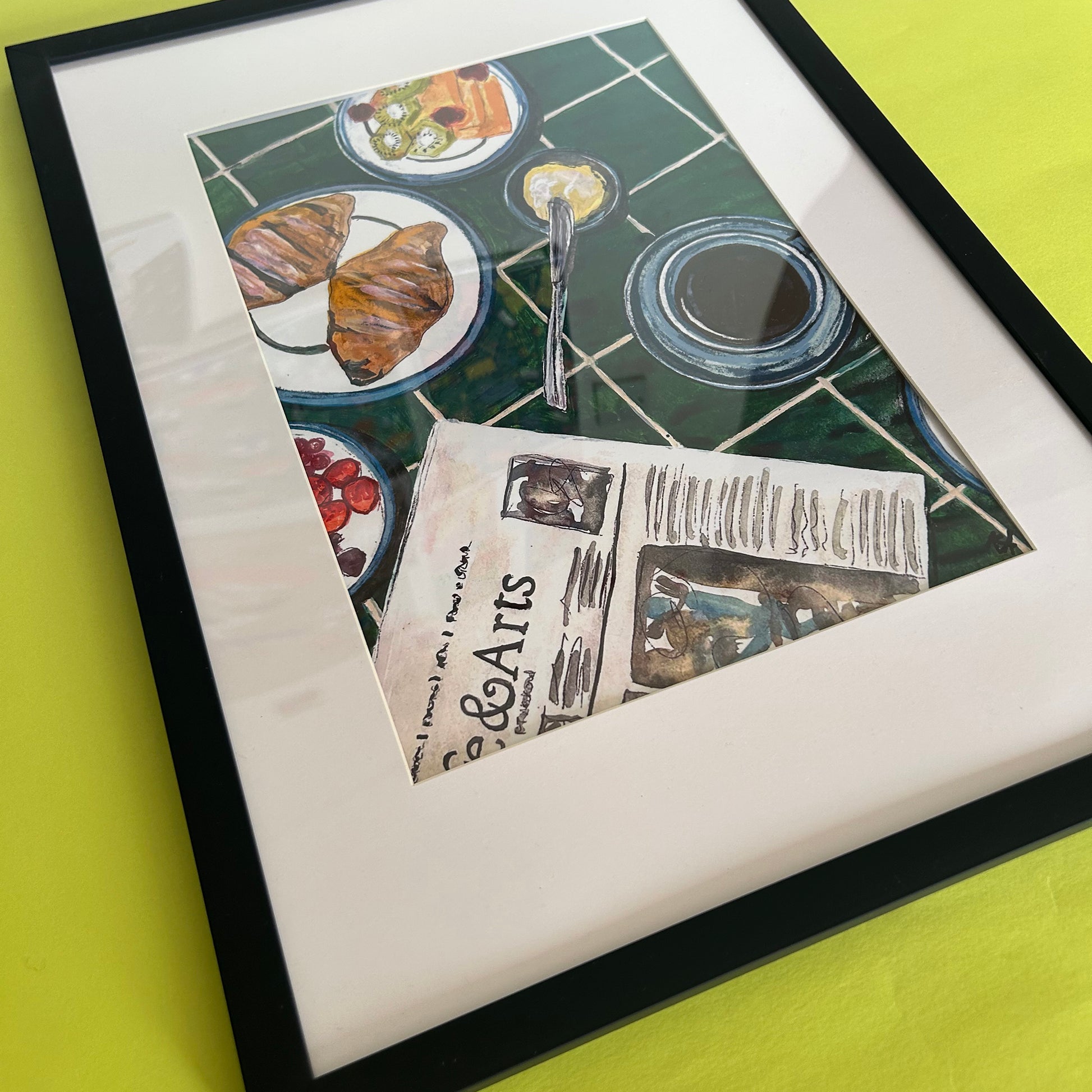 A framed art print featuring a still life illustration of a breakfast table with croissants, plates, a newspaper, and a glass of milk on a green tiled background, displayed on a bright yellow surface.