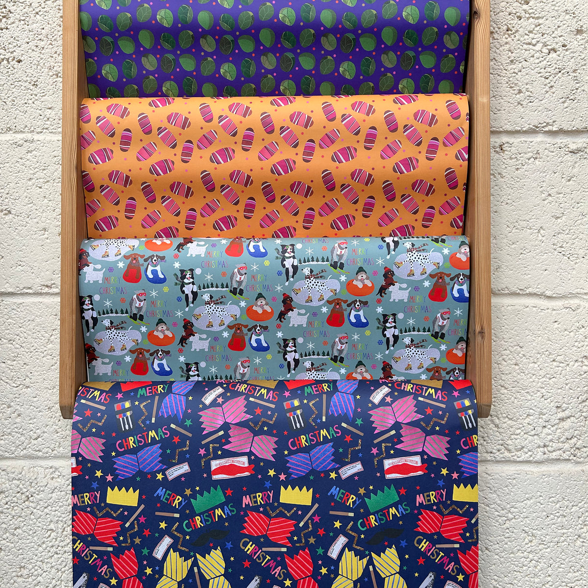 Four rolls of Christmas-themed wrapping paper displayed vertically in a wooden rack against a white painted brick wall. The top roll has green and purple sprout motifs, the second is yellow with pigs in blankets, the third features a light blue and white winter dogs pattern, and the bottom roll is dark blue with brightly coloured Christmas crackers. 