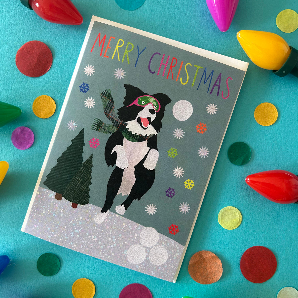 A Christmas greeting card featuring a black and white dog wearing a green hat and scarf, standing in snow with a tree and the words 'MERRY CHRISTMAS' at the top. The card is photographed from above on a bright turquoise surface surrounded by colourful craft lights and confetti.