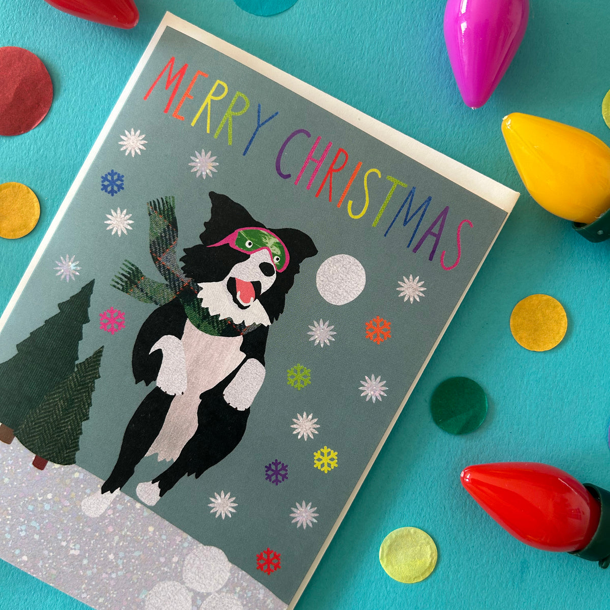 A Christmas greeting card featuring a black and white dog wearing pink goggles and a green scarf, with colourful 'MERRY CHRISTMAS' text on a teal background. The card is styled in a flat-lay with colourful light bulbs and confetti on a bright turquoise surface.