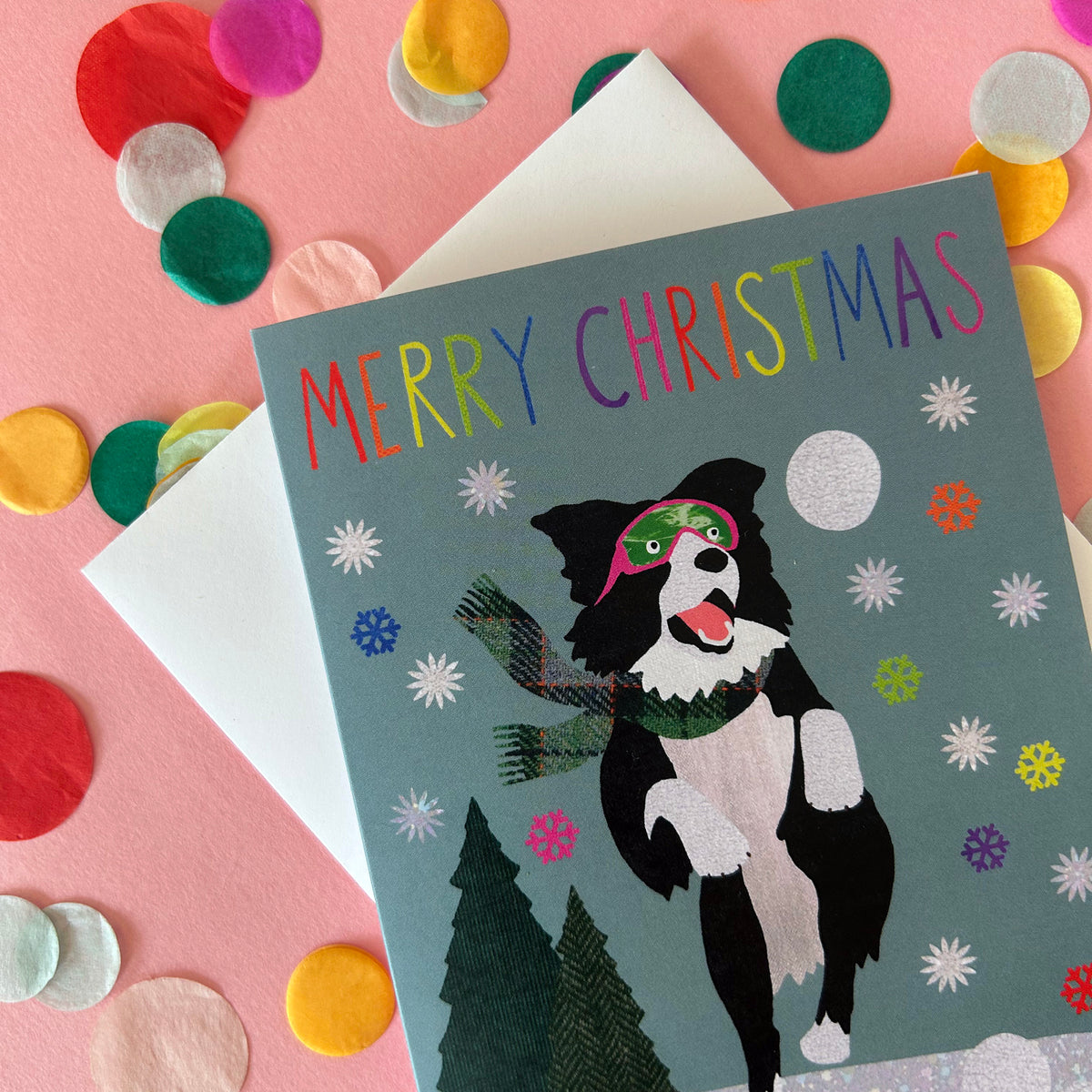 A Christmas greeting card featuring a black-and-white dog wearing a green hat and scarf on a muted blue-grey background, with the words “MERRY CHRISTMAS” in rainbow letters. The card is styled on a pink surface with a white envelope behind it and colourful felt confetti scattered around.