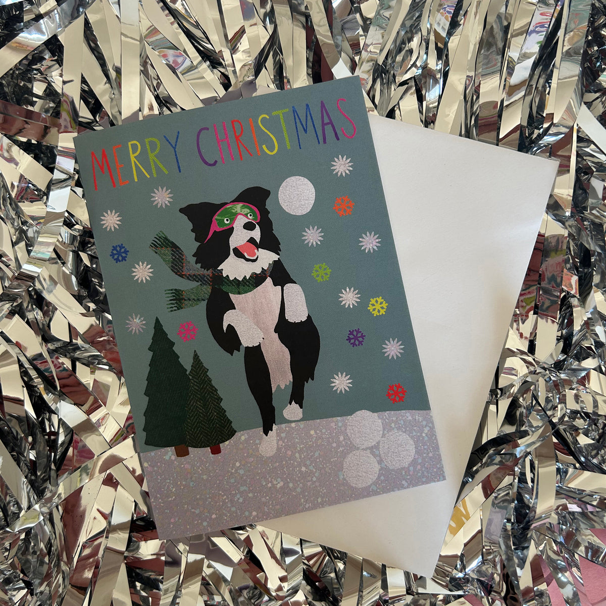 A Christmas greeting card featuring a black and white dog dressed as Santa with green goggles, surrounded by snowflakes, a tree, and the text 'MERRY CHRISTMAS' in rainbow letters. The card is shown with its white envelope on a background of shiny silver tinsel.