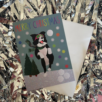A Christmas greeting card featuring a black and white dog dressed as Santa with green goggles, surrounded by snowflakes, a tree, and the text 'MERRY CHRISTMAS' in rainbow letters. The card is shown with its white envelope on a background of shiny silver tinsel.