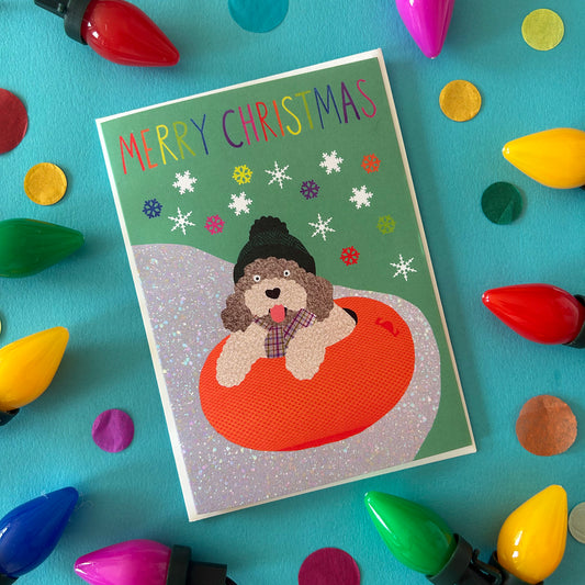 A Christmas greeting card featuring a cockapoo wearing a beanie and plaid scarf, sitting in an orange inflatable ring on a green snowy background with colourful snowflakes and the text 'MERRY CHRISTMAS'. The card is styled in a flat-lay on a bright teal surface surrounded by colourful Christmas light bulbs and confetti.