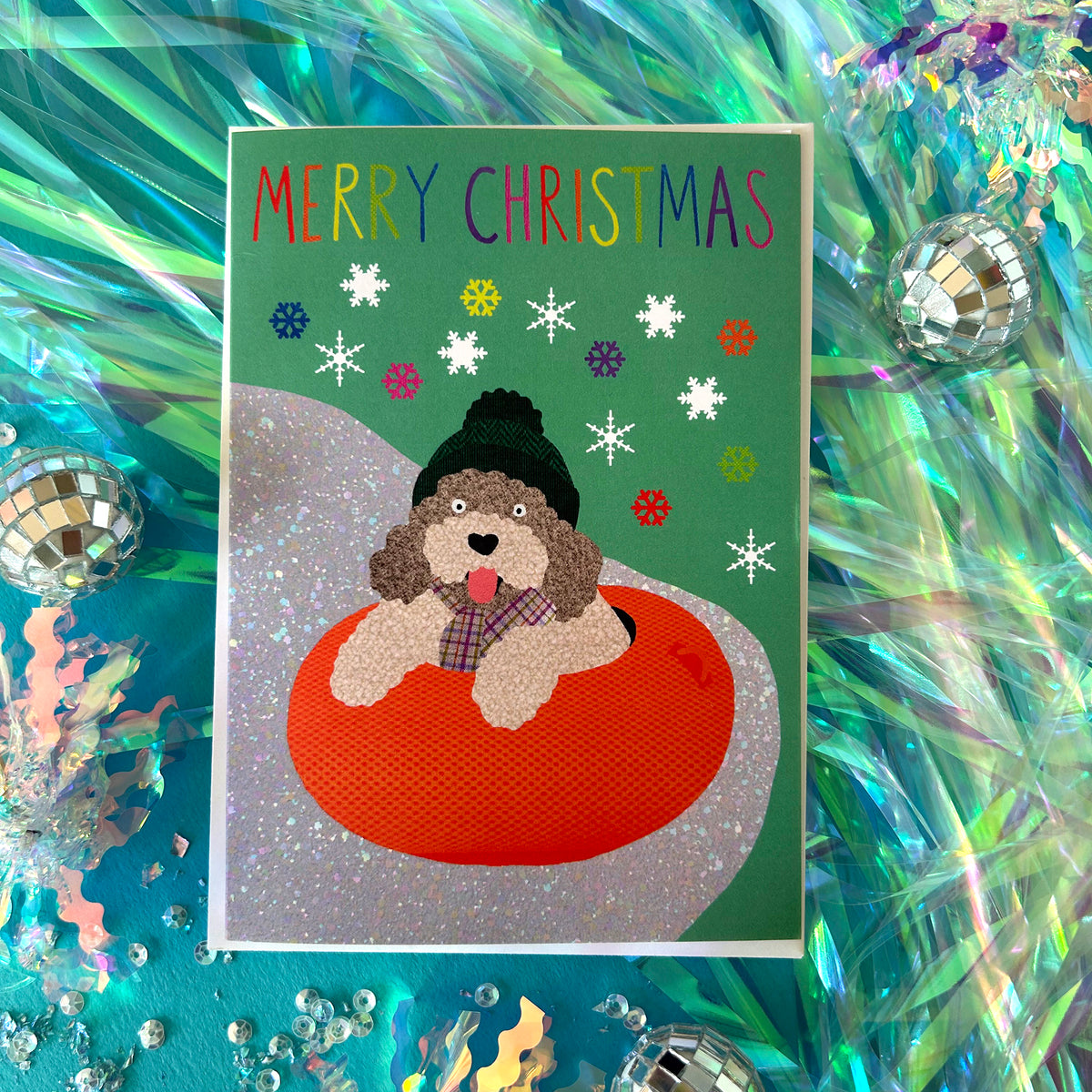 A Christmas greeting card featuring a cockapoo sitting in an orange inner tube on a green background with colourful snowflakes and the text 'MERRY CHRISTMAS'. The card is displayed on a festive, iridescent teal background decorated with disco balls and confetti.