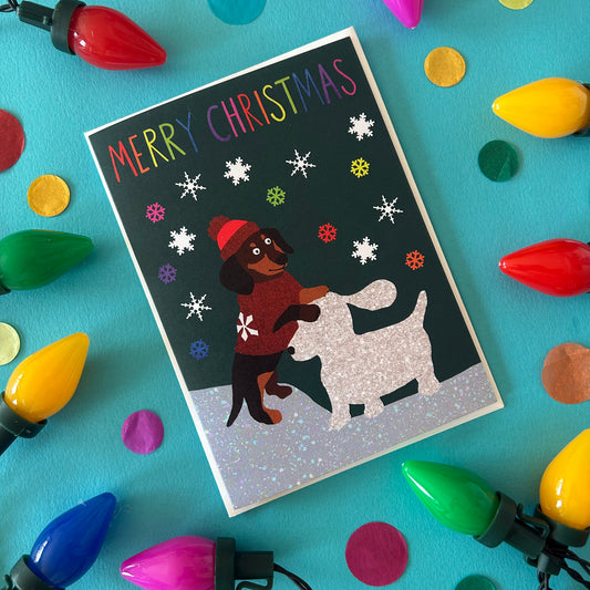 A Christmas greeting card featuring a dachshund in a red hat and sweater building a snow dog, surrounded by colourful Christmas lights and confetti on a turquoise background.
