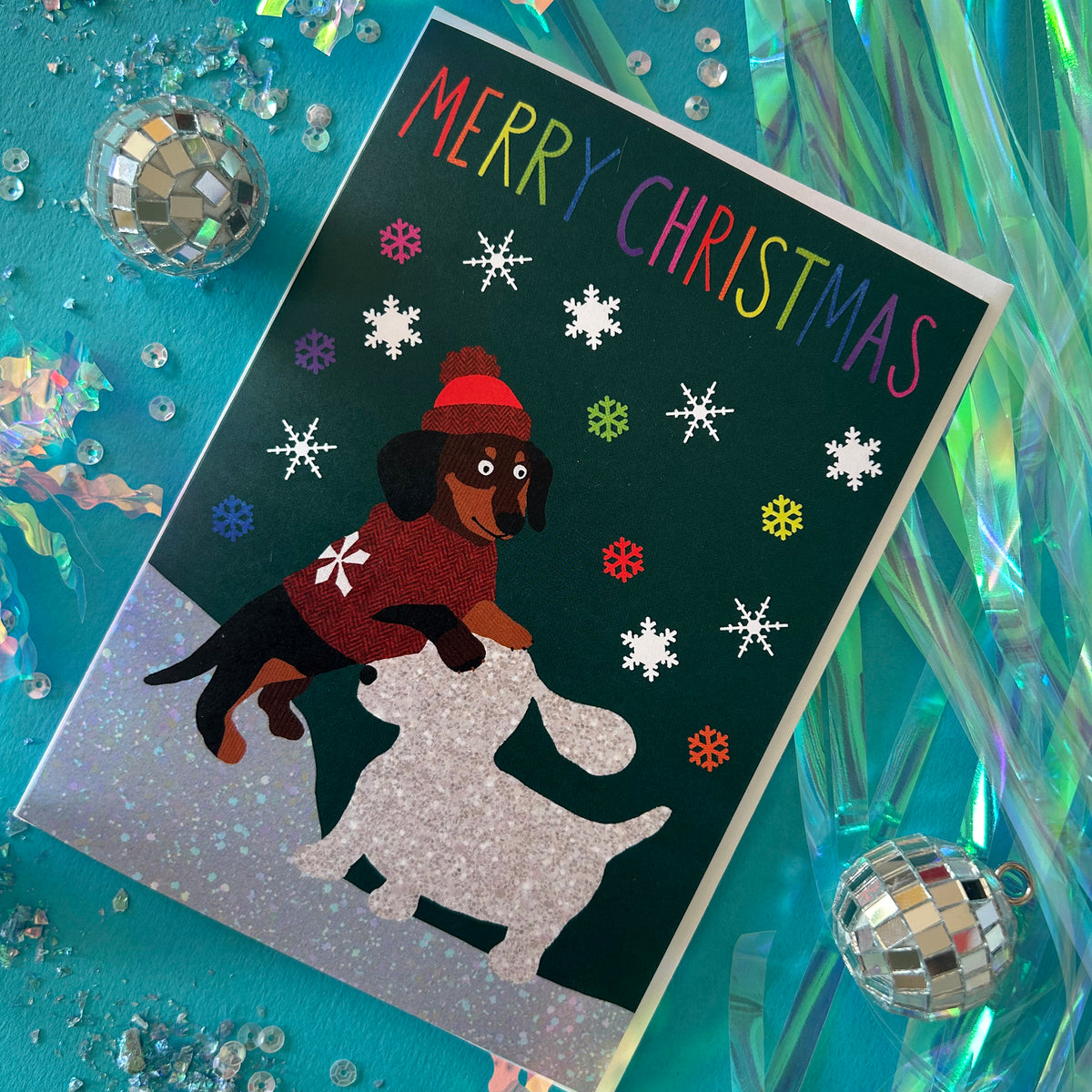 A Christmas greeting card featuring a dachshund wearing a red sweater and hat building a snow dog, surrounded by snowflakes and the text 'MERRY CHRISTMAS' in rainbow letters. The card is displayed at an angle on a turquoise background with holiday decorations, including disco balls and iridescent foil strips.