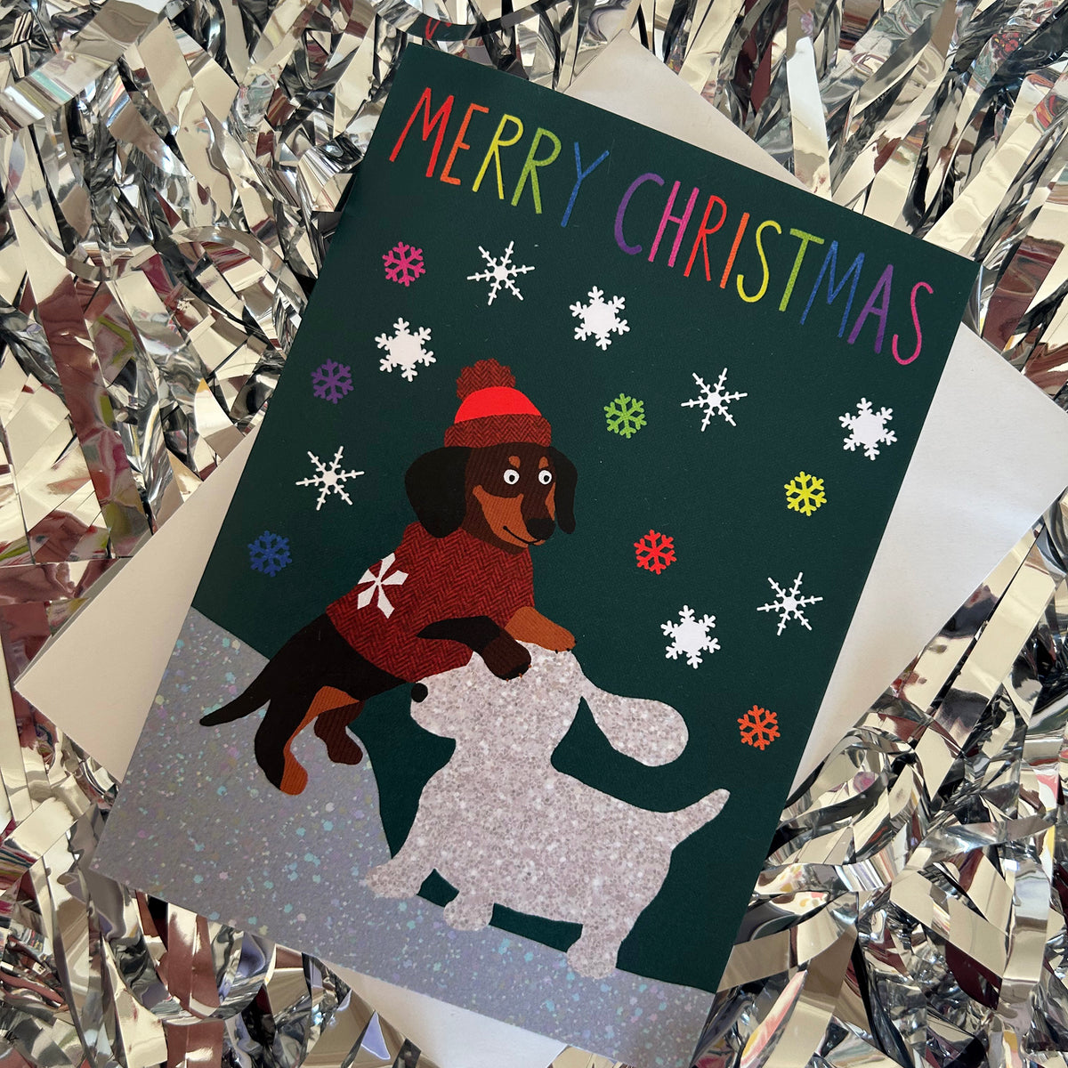 A Christmas greeting card featuring a dachshund in a red sweater and hat building a snow dog, with 'MERRY CHRISTMAS' in colourful lettering. The card is placed on top of a white envelope amidst shiny silver tinsel.