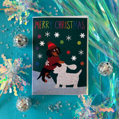 A Christmas greeting card featuring a dachshund in a red sweater and hat building a snow dog, surrounded by festive confetti, tinsel, and mini disco balls on a bright teal background.