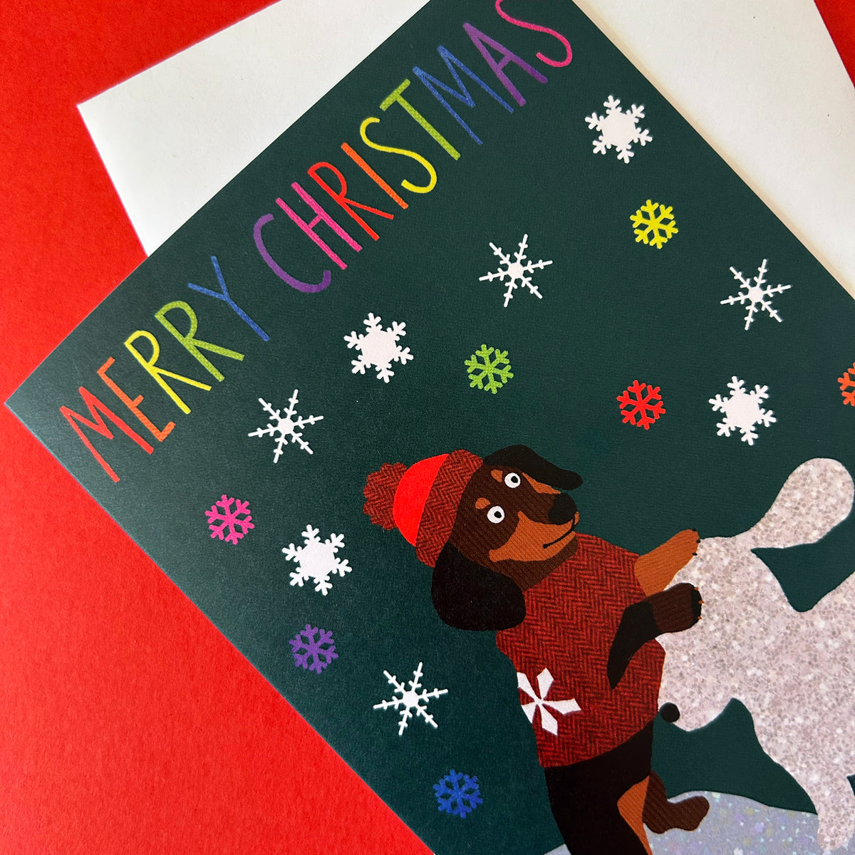 A Christmas greeting card featuring the text 'MERRY CHRISTMAS' in rainbow letters, decorated with white snowflakes on a dark green background. A dachshund wearing a red hat and red sweater is shown on a silver glittery surface.